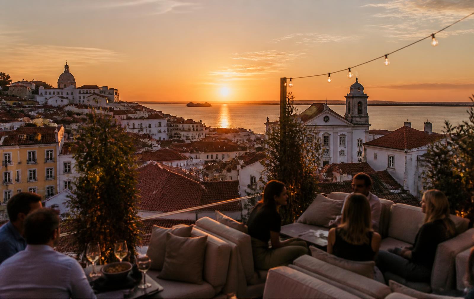 A rooftop bar terrace in Lisbon at sunset, with views over the city's red roofs toward the river